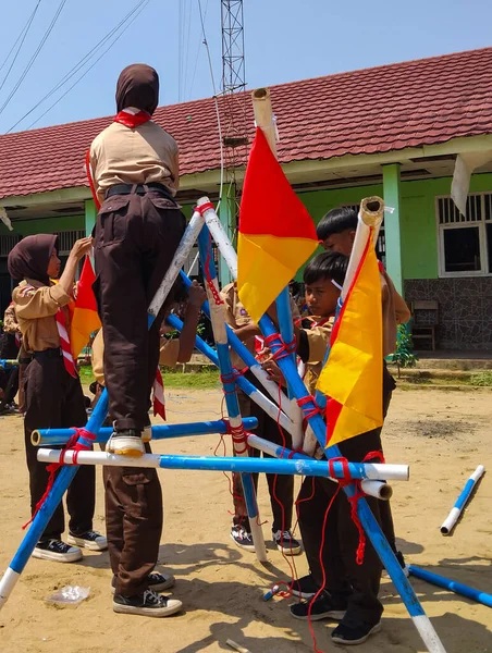 Bir grup Endonezyalı izci, kırmızı iplerle bağlanmış mavi ve beyaz direklerle bir yapı inşa etmek için açık havada birlikte çalışıyorlar.