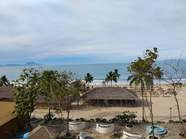 Palmiye ağaçları, bambu kulübeleri ve bulutlu bir gökyüzünün altında hafif okyanus dalgaları olan sakin bir tropikal plaj sahnesi.