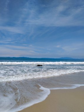 Yumuşak okyanus dalgalarının berrak mavi gökyüzünün altında kumlu bir sahile vuruşunun huzurlu görüntüsü.