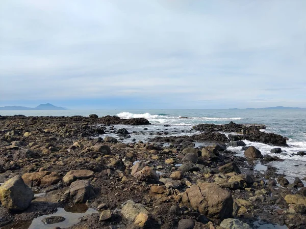 Kıyı şeridi boyunca dağılmış karanlık volkanik kayaların olduğu sakin bir sahil manzarası. Yumuşak okyanus dalgaları, parlak mavi gökyüzünün altında onlara çarpıyor.