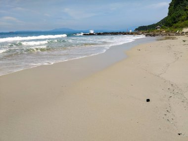 Yumuşak okyanus dalgalarının berrak mavi gökyüzünün altında kumlu bir sahile vuruşunun huzurlu görüntüsü.