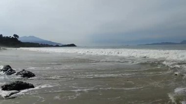 Okyanus dalgalarının sakin manzarası açık mavi gökyüzünün altındaki kumlu bir sahile hafifçe çarpıyor.