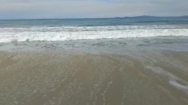 Okyanus dalgalarının sakin manzarası açık mavi bir gökyüzünün altındaki kumlu bir sahile nazikçe çarpıyor.