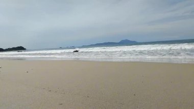 Okyanus dalgalarının sakin manzarası açık mavi bir gökyüzünün altındaki kumlu bir sahile nazikçe çarpıyor.