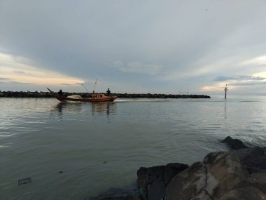 Lampung, Endonezya - 20 Kasım 2025: Küçük bir ahşap balıkçı teknesi huzurlu ve bulutlu bir günbatımında sakin kıyı sularında süzülüyor