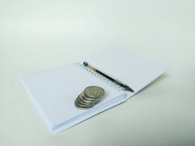 Açık bir spiral deftere yerleştirilmiş gümüş para yığını. Temiz beyaz arka planda tükenmez kalemle birlikte.