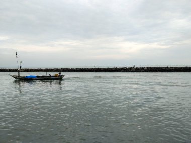 LAMPUNG, INDONEZYA - 10 Aralık 2025: Balıkçıları taşıyan küçük bir balıkçı teknesi taştan bir dalgakıranın yanındaki sakin kıyı sularında süzülüyor