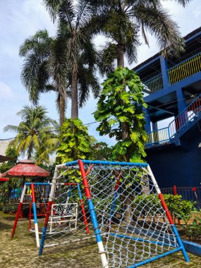 Uzun tropikal palmiye ağaçları ve yemyeşil bitkilerle çevrili renkli bir açık hava oyun alanı.