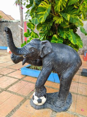 Bir futbol topunu tropik bir bahçede ayağıyla dengeleyen neşeli bir fil heykeli.