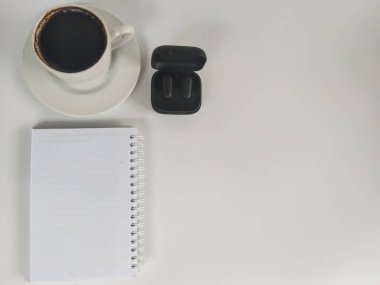 Minimalist düz fotograf uzerinde bir fincan sade kahve, acik siyah kablosuz kulaklik kutusu ve bos bir spiral defter var.