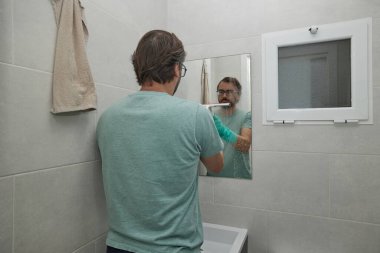 Banyodaki aynayı sprey ve süngerle temizleyen adam yeşil eldiven giyiyor. Ev işleriyle uğraşan ve evde temizlik yapan biri. Hijyen, temizlik ürünleri ve günlük rutin