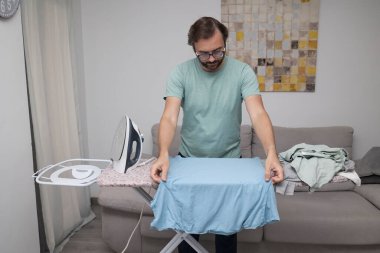 Adam evde giysileri ütülüyor. Çamaşır yıkamak için ütü tahtası kullanan biri. Birlikte ev işi yapan bir çift. Ev temizliği, ev işleri ve günlük rutin işler kavramı.