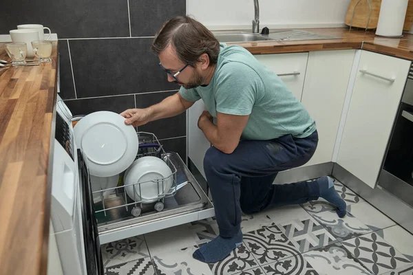 Modern mutfakta bulaşık makinesini yükleyen adam. Ev işleri yapan, bulaşık yıkayan ve evde mutfak gereçleri düzenleyen biri. Ev hayatı kavramı, ev temizliği ve modern yaşam tarzı..