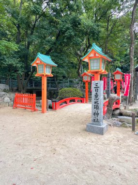 Japonya 'da yemyeşil ağaçlarla çevrili bir Shinto tapınağının girişindeki parlak kırmızı köprü ve fenerler. Fukuoka, Japonya - 31 Ekim 2025.