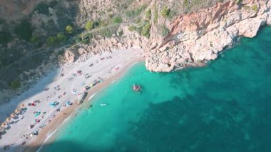 İspanya 'nın Benitatxell kentindeki Cala del Moraig sahilinin tepeden aşağı insansız hava aracı görüntüsü turkuaz Akdeniz suları, uçurumlar ve sahilde güneşlenen turistler..