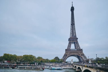 Seine Nehri üzerinde yükselen Eyfel Kulesi bulutlu bir sonbahar gününde yakınlardan geçen teknelerle. Paris, Fransa - 10 Ekim 2025.
