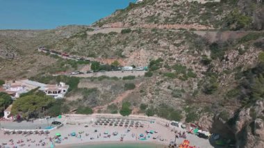 Cala Granadella plajının insansız hava aracı görüntüsü uçurumlardaki virajlı yol boyunca park edilmiş arabalar ve kumlu sahilde dinlenen insanlar. Javea, İspanya - 4 Ekim 2025.