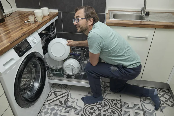 Modern mutfakta bulaşık makinesini yükleyen adam. Ev işleri yapan, bulaşık yıkayan ve evde mutfak gereçleri düzenleyen biri. Ev hayatı kavramı, ev temizliği ve modern yaşam tarzı..