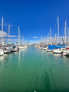 Turkuaz sudaki yelkenli tekneleriyle dolu bir marinanın panoramik görüntüsü, kıyı yaşam tarzını ve deniz atmosferini yakalıyor. Valencia, İspanya - 8 Kasım 2025.