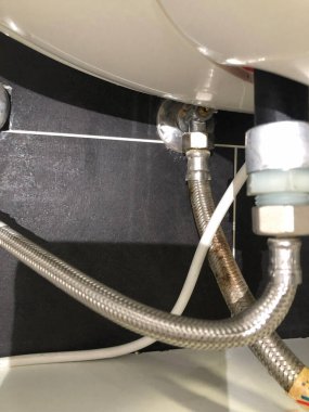 Banyo lavabosunun altındaki su ısıtıcı borularının ve tesisat bağlantılarının yakından görüntüsü bakım ve teftiş sırasında metal örgülü hortumları, tesisatları ve su tedarik hatlarını gösteriyor..