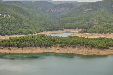 Dağ yamaçlarıyla çevrili turkuaz göl sularına uzanan orman yarımadasının havadan görünüşü.