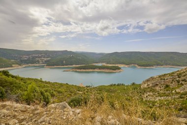 Ormanlık tepeler ve kumlu kıyılarla çevrili bir dağ gölünün geniş manzarası yumuşak mavi bir gökyüzünün altında.