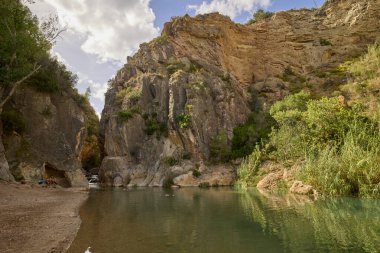Parlak mavi gökyüzünün altında yeşil bitki örtüsünün üzerinde yükselen katmanlı kayalık kanyon duvarlarının görüntüsü.