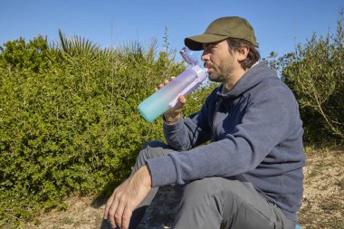 Güneşli bir günde, açık hava yürüyüşü sırasında yerde oturup bir spor şişesinden su içen bir adam. Çevrede yeşil çalılar ve doğal manzara.