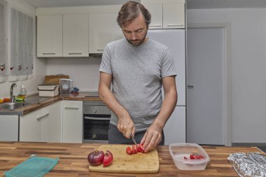 Bir adam mutfakta taze malzemeler hazırlarken ahşap bir tahtada olgun domatesler kesiyor..