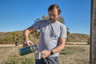 Kırsal piknik masasında termostan metal bardağa sıcak içecek döken bir adam..