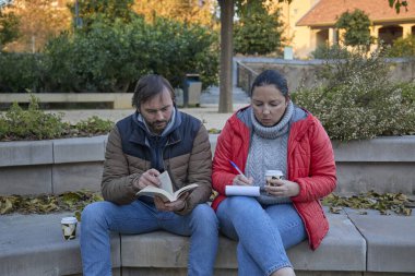Bir adam kitap okuyor ve bir kadın bir sonbahar parkında yan yana otururken deftere yazı yazıyor..