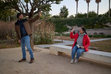 Bir adam bankta kitap okuyan bir kadına el sallıyor..