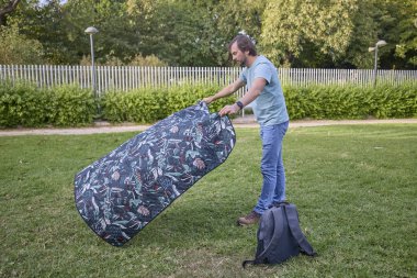 Orta yaşlı bir adam piknik yeri hazırlıyor. Güneşli yeşil bir parktaki çimlere örtü seriyor..
