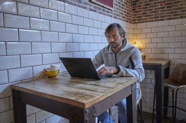 Sade bir kafedeki ahşap bir masada oturan adam dizüstü bilgisayar kullanıyor, etrafı beyaz fayanslarla çevrili ve sıcak dekoratif ışıklandırmayla çevrili..