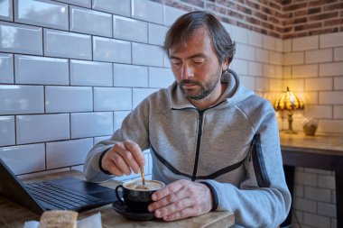 Gri kapüşonlu bir adam kafenin ahşap masasında oturuyor, dizüstü bilgisayarla çalışırken bir fincan kahve karıştırıyor..