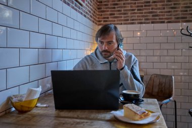 Kafede dizüstü bilgisayarla çalışan bir adam akıllı telefondan konuşuyor..
