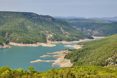 Parçalı bulutlu bir gökyüzünün altında İspanya 'da yeşil orman ve tepelerle çevrili bir turkuaz dağ gölü geniş panoramik manzarası. Huzurlu doğal manzara ve vahşi doğa.