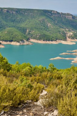 Parçalı bulutlu bir gökyüzünün altında İspanya 'da yeşil orman ve tepelerle çevrili bir turkuaz dağ gölü geniş panoramik manzarası. Huzurlu doğal manzara ve vahşi doğa.