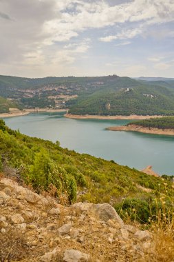 Çam ormanları ve bulutlu gökyüzünün altındaki kayalık dağ yamaçlarıyla çevrili bir rezervuarın panoramik görüntüsü.
