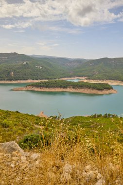 Ormanlık tepeler ve kumlu kıyılarla çevrili bir dağ gölünün geniş manzarası yumuşak mavi bir gökyüzünün altında.