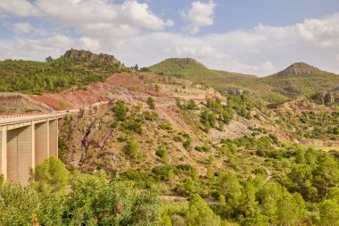 Kırmızı kaya tabakası, çam ağaçları ve parlak yaz ışığı ile sarp bir dağ vadisini kaplayan yüksek beton köprü..