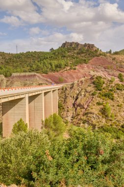 Kırmızı kaya tabakası, çam ağaçları ve parlak yaz ışığı ile sarp bir dağ vadisini kaplayan yüksek beton köprü..
