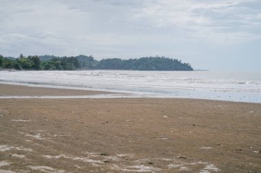 Yumuşak deniz dalgaları, kumlu kıyı şeridi ve bulutlu bir gökyüzünün altındaki uzak bir ormanlık adanın geniş manzarası..