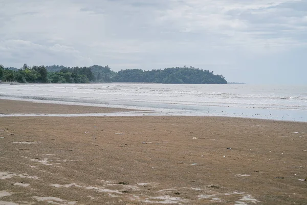 Yumuşak deniz dalgaları, kumlu kıyı şeridi ve bulutlu bir gökyüzünün altındaki uzak bir ormanlık adanın geniş manzarası..