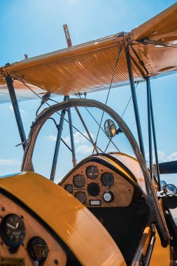 Sarı renkli çift kanatlı bir uçağın kokpit manzarası ahşap enstrüman panelini gösterirken göstergeler, kontrol detayları ve destekleyici destekleyici destekler. Açı yukarı, kumaşla kaplı kanada doğru bakıyor. Güneş ışığı, dokuyu parlak bir kanada vurguluyor.