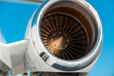 Türbin bıçaklarını ve metal yüzeyleri gösteren bir jet motoru beşiğinin yakın çekimi. Parlak konut ışığı yansıtıyor, emme vantilatörü mavi gökyüzü arka planında açıkça görülüyor..
