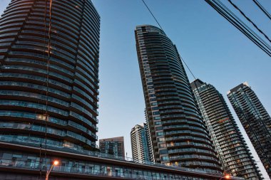 Sıcak akşam ışığında şehir siluetine yerleşik kuleler hakim. Düşük açı perspektifi ölçek ve derinliği artırırken, sokak lambaları ve kablolar gibi kentsel detaylar içerik ekler. Alacakaranlık 'ın serin tonları modern geometri ve yansıtıcı cam sörfünü dengeler.