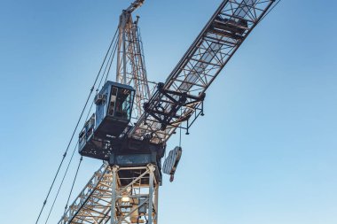 Bir kule vincinin yakın çekimi çelik iskeletini, kablolarını ve santral kabinini temiz bir gökyüzüne karşı yakalar. Düşük bir açıdan çekilen görüntü endüstriyel tasarım ve gücü vurguluyor. Temiz bileşim ve açık arkaplan kopyalama alanı