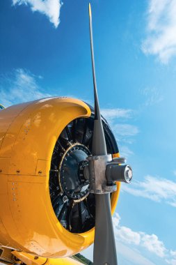 Klasik sarı uçak radyal motorunun düşük açılı yakın çekimi ve önden gümüş pervane. Motor kaplaması açık, ayrıntılı radyal motor silindirlerini gösteriyor. Yerden çekilen beyaz bulutlu parlak mavi gökyüzüne karşı.