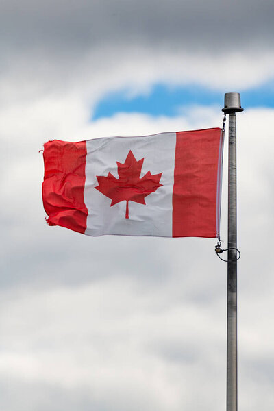 Канадский флаг с красным кленовым листом, размахивающим горизонтально на металлическом флагштоке против серого облачного неба. Маленький участок голубого неба, видимый сквозь облака на заднем плане.
